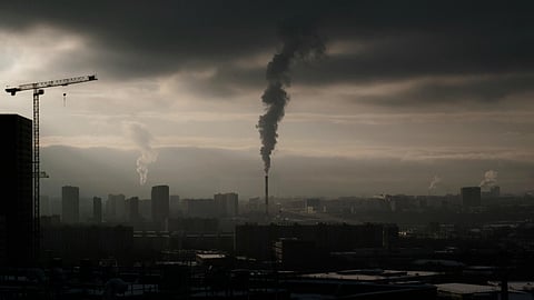 A dark city skyline with smokestacks releasing thick smoke under cloudy skies.