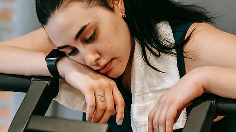 A tired woman leaning on a treadmill with a towel around her neck and a smartwatch.