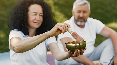 A woman gently plays a singing bowl with a mallet outdoors, while a man sits in the background. 