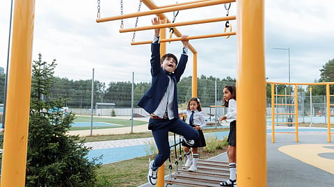Three children in school uniforms play on yellow playground monkey bars. 