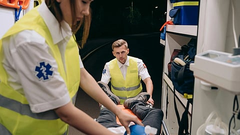 Two paramedics in yellow vests assist a patient on a stretcher inside an ambulance at night. 