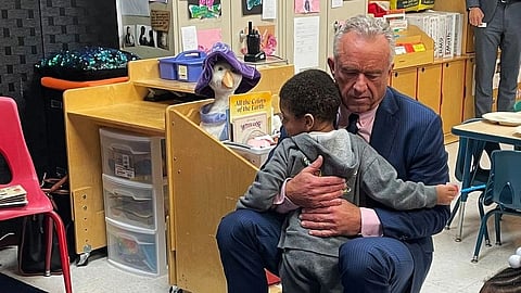 A man in a suit kneels to comfort a child in a classroom, offering a warm and reassuring embrace.
