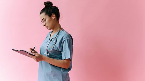 A healthcare worker in scrubs with a stethoscope writes on a clipboard against a bright pink background.