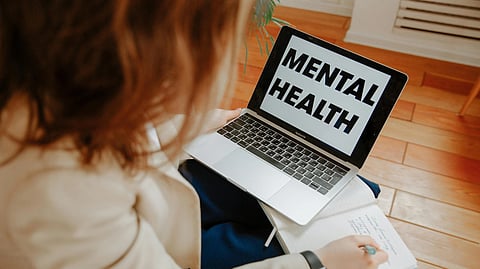 A person holds a notebook beside a laptop screen displaying “MENTAL HEALTH.”