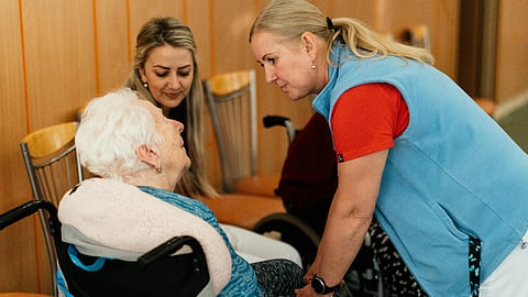 Caregiver in blue vest speaks with elderly woman in a wheelchair.