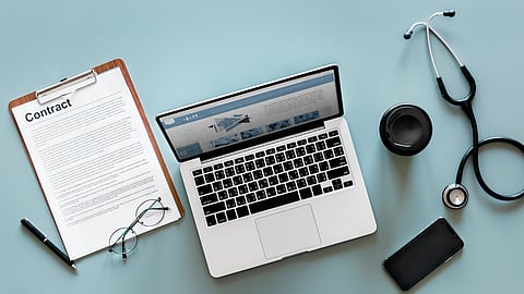 Laptop with a medical website sits beside a stethoscope, phone, glasses, and documents on a desk.