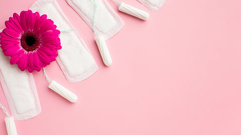 White sanitary pads and tampons are arranged diagonally on a pink background with a bright pink gerbera daisy.