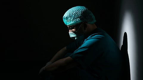 A healthcare professional in surgical scrubs and cap sits alone in a darkened space, head bowed, representing the silent mental health crisis among resident doctors in India.