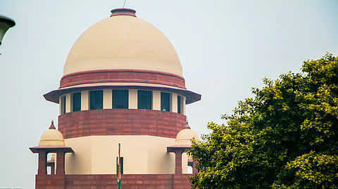 The Supreme Court of India building with its dome, national flag, and green trees under a clear sky.