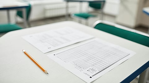 Test papers and a pencil on a classroom desk with blurred chairs in the background.