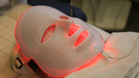 Person wearing a glowing LED facial mask emitting red light, lying on a bed.