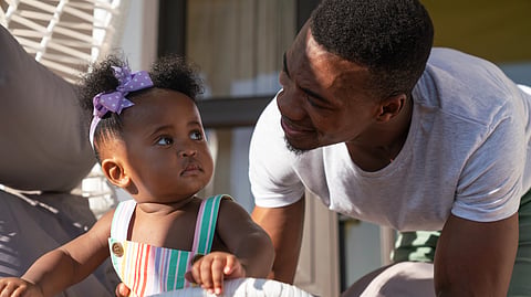 A man smiles warmly at a baby in a colorful dress with a purple bow, sitting outdoors. 