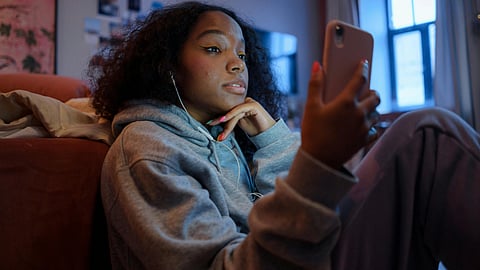 A woman in a grey hoodie sits indoors, looking at her phone with earphones in.
