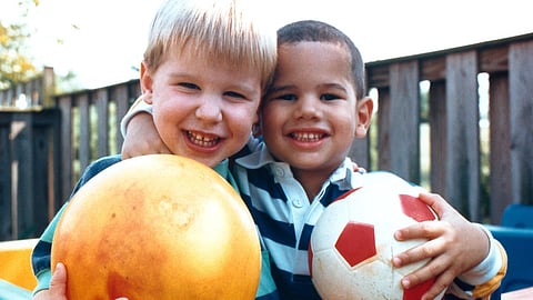 Two smiling children hug while holding a ball in a playground.