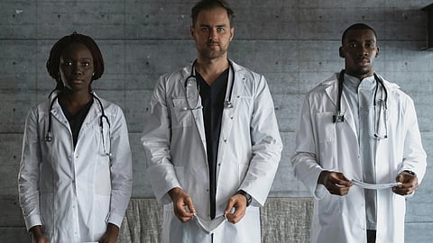 Three doctors in white coats and stethoscopes stand against a gray wall. 