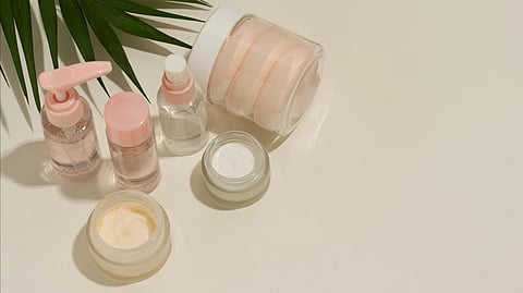 Skincare products in jars and bottles with pink lids are placed on a light surface with green leaves.