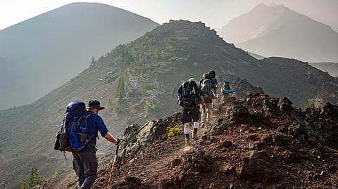 Hikers with backpacks walk on a rocky mountain ridge under a hazy sky.