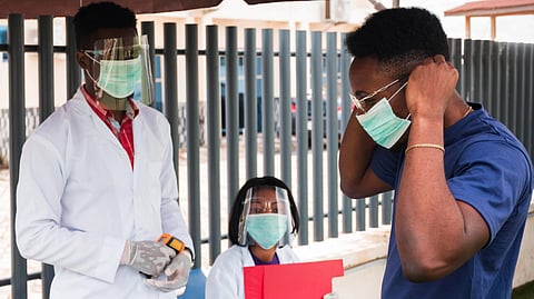 Two healthcare workers, wearing face shields and masks, assist a man adjusting his mask outdoors.