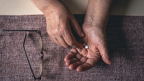 Elderly hands holding a small white pill over a purple textured mat. 