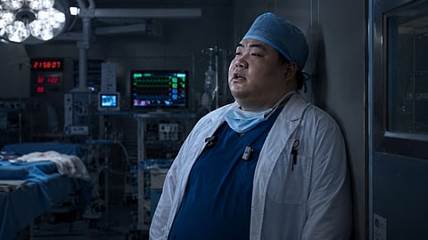 Photorealistic editorial illustration of an exhausted overweight surgeon standing alone in a dimly lit hospital operating room after a long shift, with surgical cap, loosened mask, glowing monitor lights, and visible signs of fatigue and burnout.