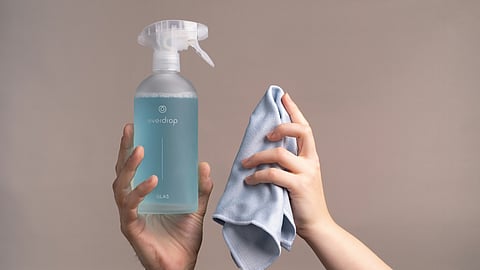 Two hands hold up a disinfectant spray bottle labeled "everdrop GLAS" with blue liquid and a light blue microfiber cloth against a plain background.