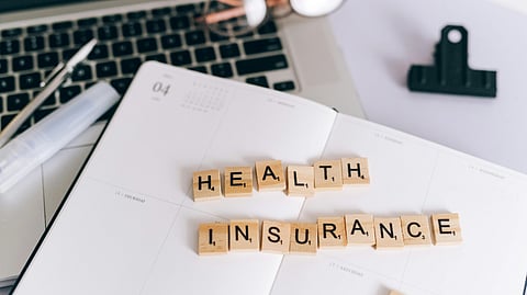 Wooden Scrabble tiles spell "Health Insurance" on an open planner atop a laptop. 
