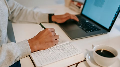 Person writing in a notebook while using a laptop beside a cup of coffee.