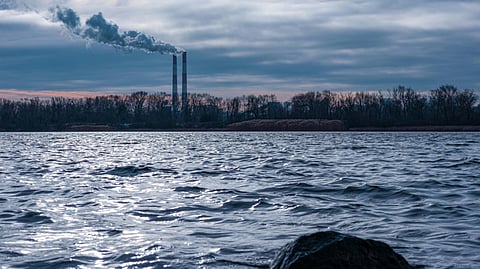 Large water body with ripples under cloudy sky, with smoke from factories and bare trees in background.