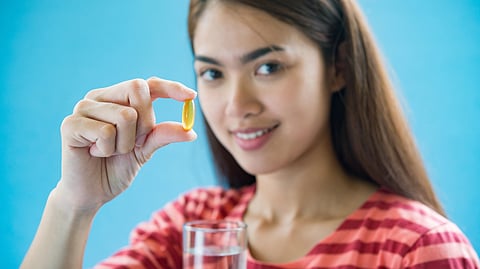 A woman in a red-striped shirt smiles while holding a yellow capsule and a glass of water.