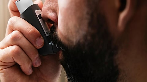 A bearded man uses an inhaler indoors to relieve his asthma symptoms.