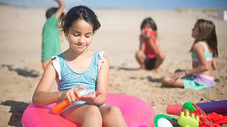 A young girl applies sunscreen on the beach while other children play in the sand behind her.