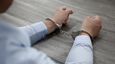 A person in a blue shirt sits at a wooden table, hands cuffed with silver handcuffs. 