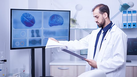 A doctor in a white coat examines a clipboard in a modern lab. A screen displays brain images.