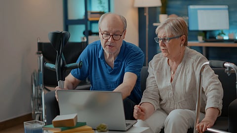 An elderly couple sits in a cozy room, focused on a laptop. 