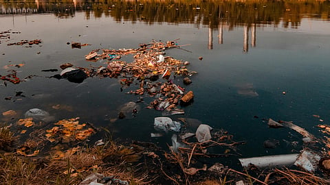 Polluted river with trash floating in the foreground, industrial chimneys and trees in the background.