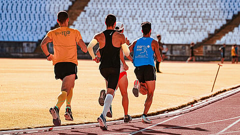 Four athletes run on a sunlit track in an empty stadium, wearing colorful athletic gear and focused expressions.