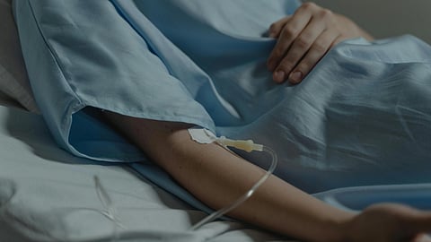 A person in a hospital bed wears a blue gown, with an IV line in their arm.