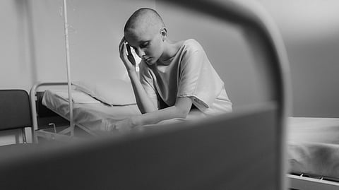 A bald person in a hospital gown sits pensively on a bed in a dim room.
