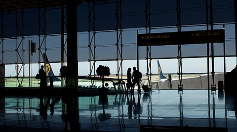 A group of people stands in an airport terminal, waiting near check-in counters and departure screens.