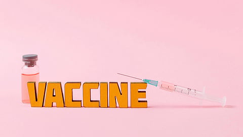 A vaccine vial and syringe placed beside bold “VACCINE” text on a pink background.