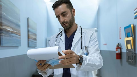 A male doctor stands in a hallway, holding a clipboard and looking focused on his tasks.