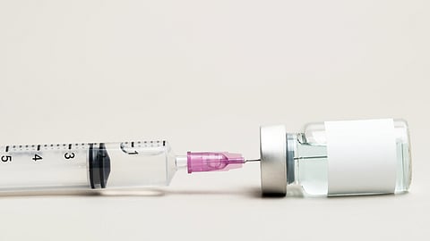 A syringe with a pink needle tip is inserted into a small glass vial of clear liquid.
