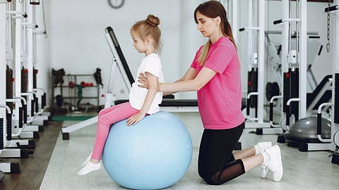 A woman kneels, supporting a young girl sitting on a blue exercise ball in a gym.