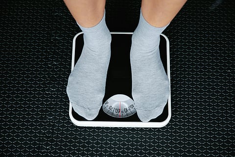 A person standing on a weighing scale viewed from above, with the analog dial showing a mid-range weight reading, representing the clinical reality that body weight and BMI are unreliable proxies for metabolic health in South Asian patients, where visceral fat accumulates at lower body weights than Western populations.