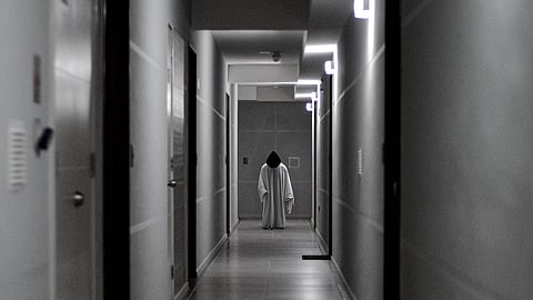 A dimly lit, monochrome hallway with a figure in a white robe standing at the end. 