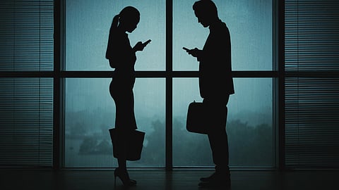 Silhouette of two business professionals standing side by side, each focused on their smartphones.