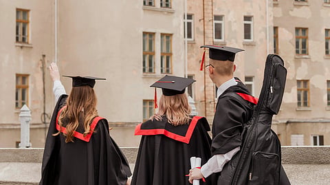 Three graduates in caps and gowns face an old building, one raising a hand.