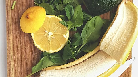 A wooden cutting board with avocado, greens, lemon slices, and a banana.