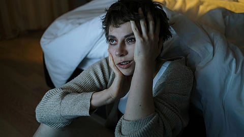 A woman sitting on a bed, resting her hands on her face, appears deep in thought or distress.