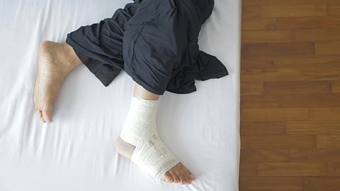 A person lying in bed, wearing dark pants, with a foot wrapped in white bandages.